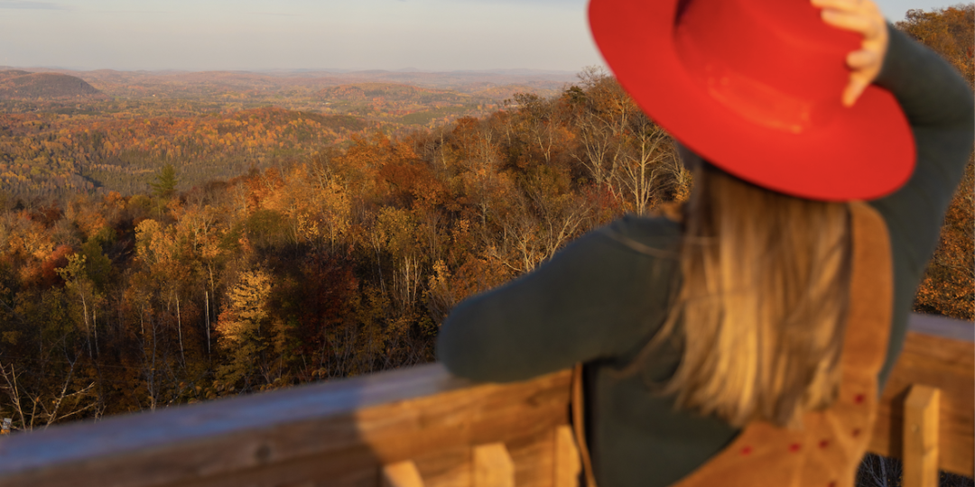 Woman in red hat and corduroy jumper standing at a wood railing overlooking fall foliage in Quebec